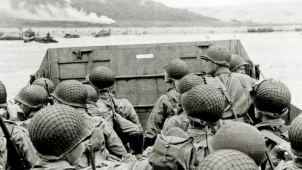 Des soldats américains aperçoivent la Normandie depuis leur bateau, avant de débarquer sur Omaha Beach. De loin, on peut apercevoir des véhicules déjà sur place et de la fumée.